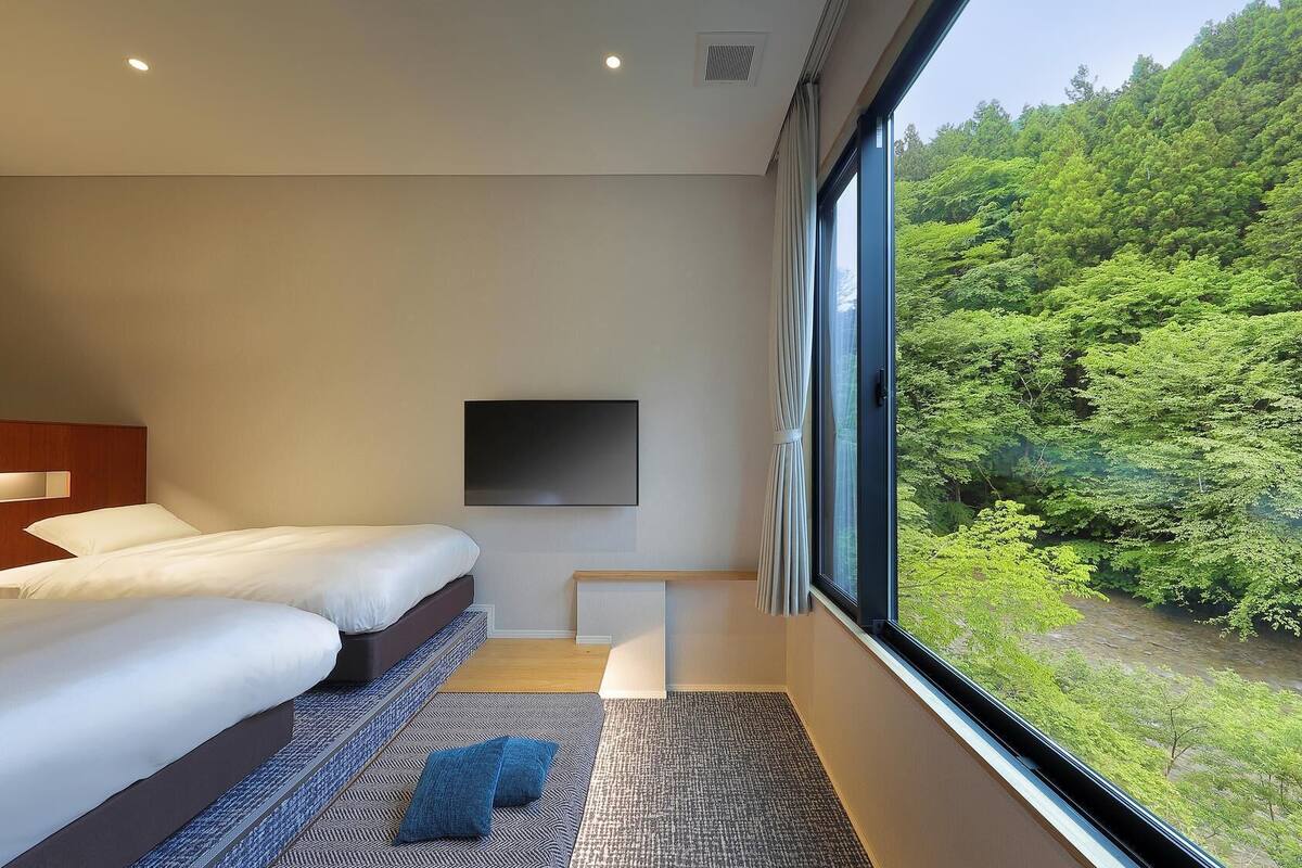 A modern hotel room with two beds, a flat-screen TV, and a large window offering a view of a forested area.