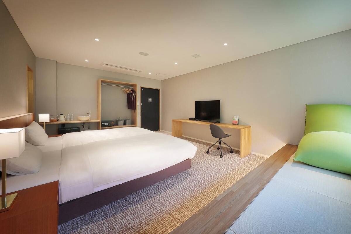A modern hotel room with a large bed, a desk with a TV, a chair, and a window with curtains.