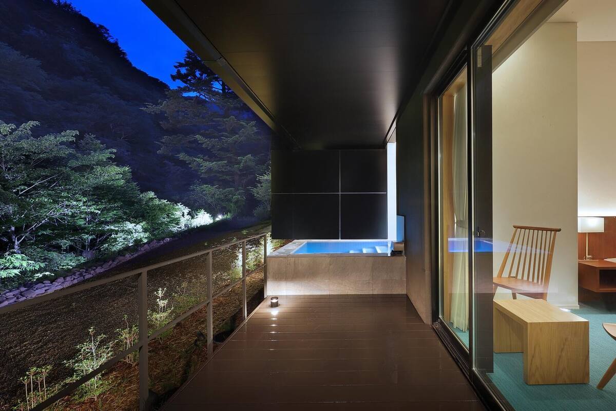 A balcony with a view of a forested area, a small pool, and a seating area with a bed and a chair.