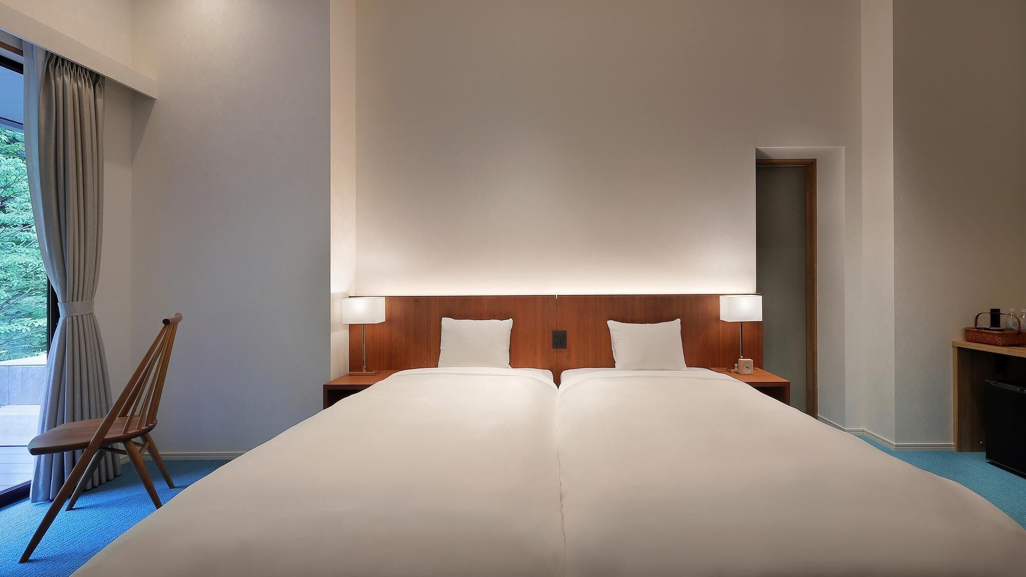 Hotellihuone, jossa on suuri sänky, kaksi yöpöytää valaisimineen, puinen sängynpääty, tuoli ja näkymä vihreälle alueelle.