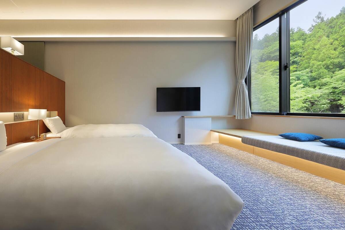 A modern hotel room with a large bed, a flat-screen TV, a seating area with cushions, and a view of a forest through a large window.