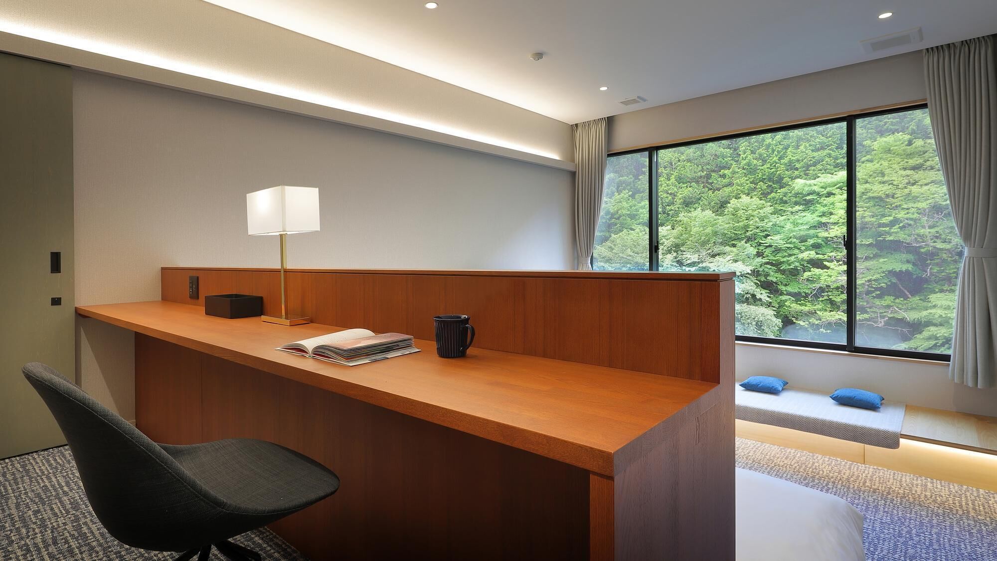 A modern office space with a wooden desk, a black office chair, a lamp, and a view of greenery through large windows.