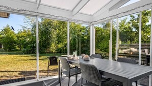 Un invernadero con una mesa y sillas de comedor, ventanas amplias y vistas a un jardín.