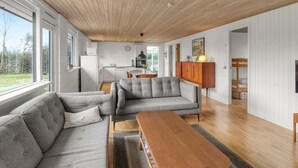 Una sala de estar moderna con techo de madera, un sofá gris, una mesa de centro de madera y una cocina al fondo.