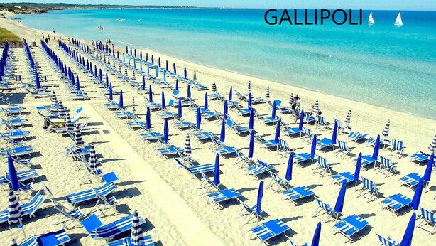 A beach with rows of blue and white striped sun loungers and umbrellas, a clear blue sea, and sailboats in the distance.