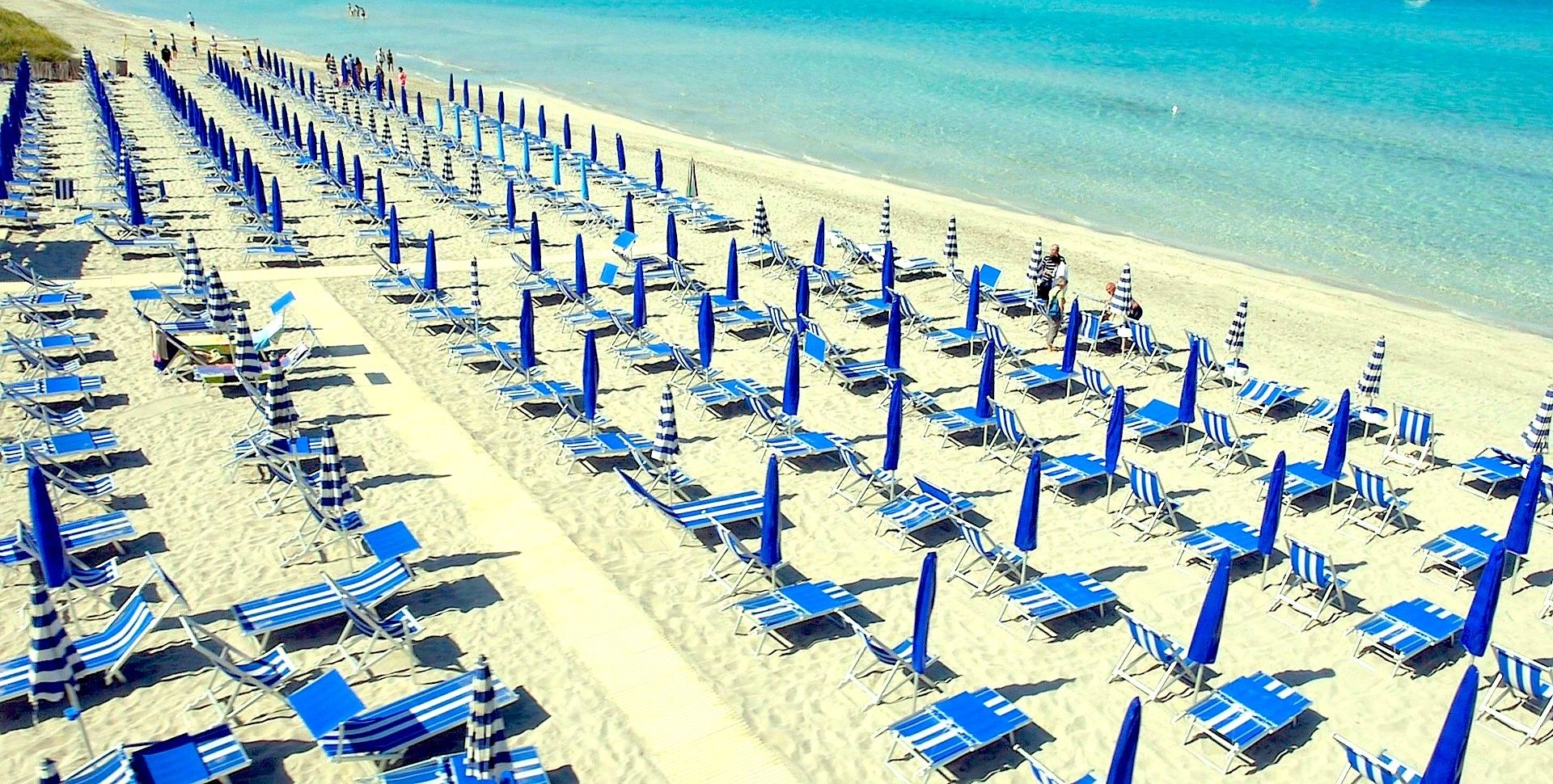 Ein Strand mit Reihen von blau-weißen Sonnenliegen und Sonnenschirmen, blauem Meer und Segelbooten in der Ferne.
