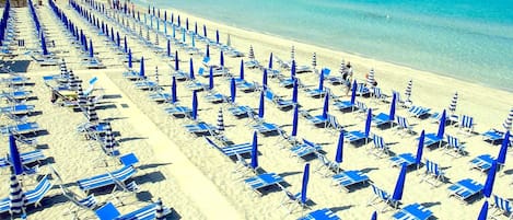 A beach with rows of blue and white striped sun loungers and umbrellas, a clear blue sea, and sailboats in the distance.