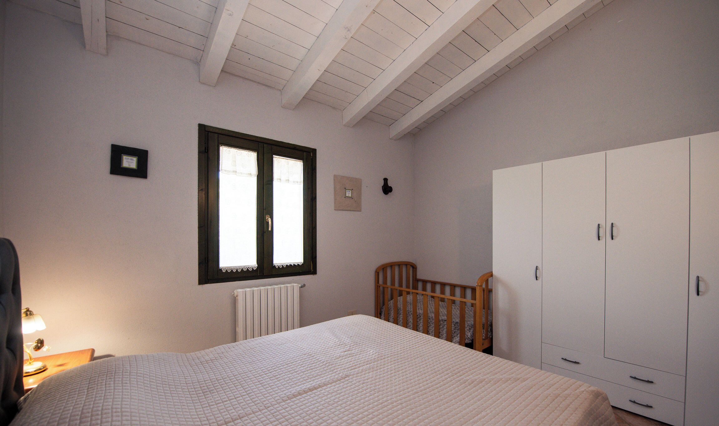 A bedroom with a bed, a wooden crib, a wardrobe, a window with curtains, and a radiator.