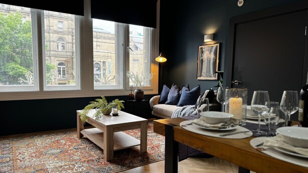 Un coin repas avec une table dressée pour deux personnes, un canapé avec des coussins, un tapis et une vue sur un immeuble à travers de grandes fenêtres.
