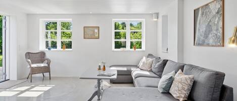 Una sala de estar moderna con un sofá seccional gris, un sillón de mimbre y una mesa de centro. Hay ventanas grandes con vistas a la vegetación.