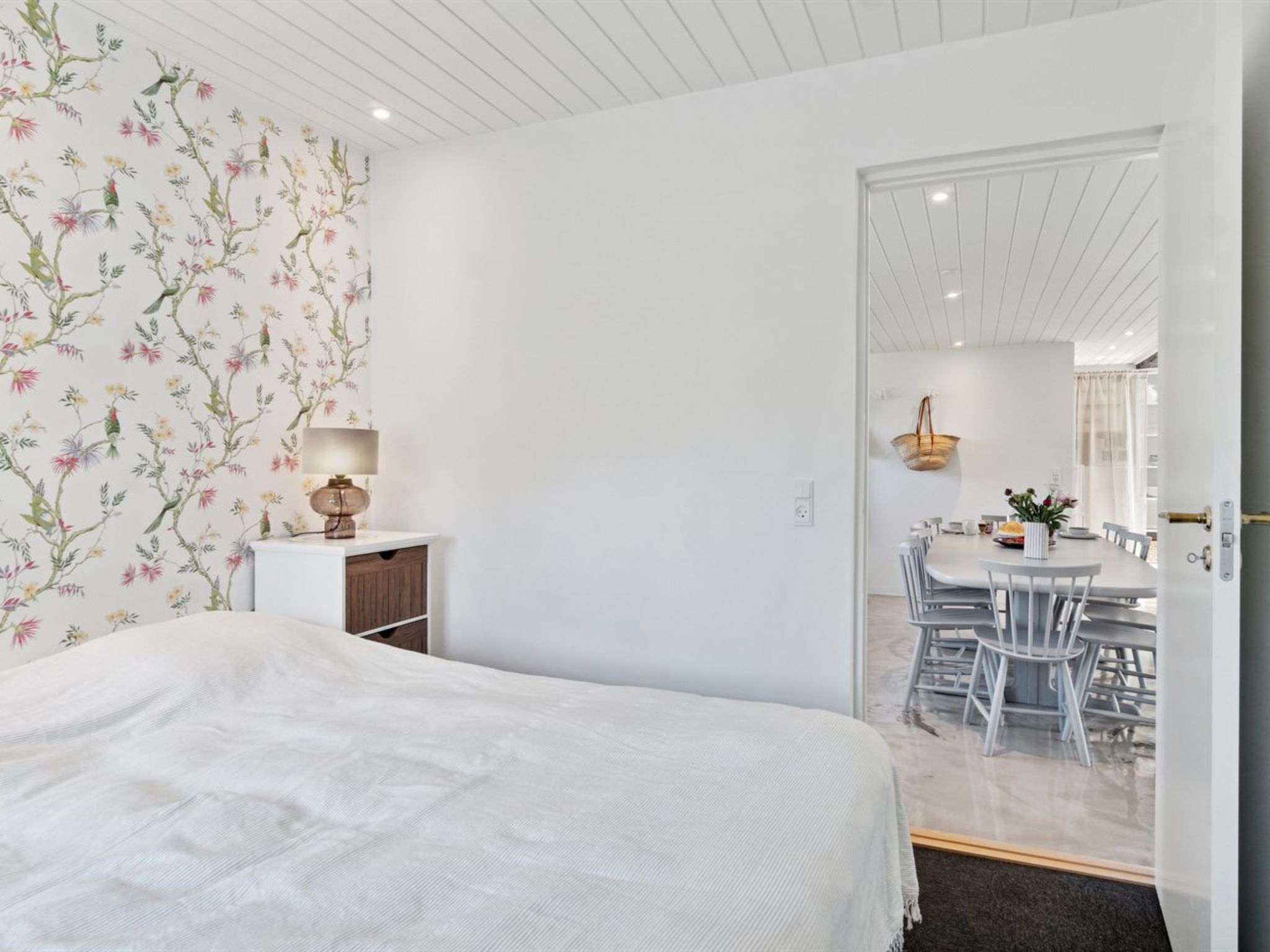 Une chambre à coucher comprenant un lit, une table de chevet avec une lampe et une armoire. Une porte donne accès à un coin repas avec une table et des chaises.