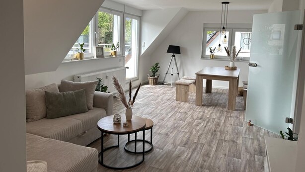 A modern loft apartment with a sloped ceiling, large windows, a dining area, and a cozy seating area with a sofa and coffee table.