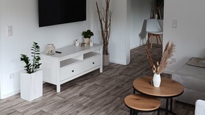 A modern living room with a flat-screen TV mounted on the wall, a white console table with plants, a round wooden coffee table, and a grey sofa.