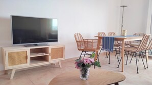 A modern living room with a flat-screen TV on a wooden cabinet, wicker chairs around a dining table, and a vase with flowers on a wooden table.