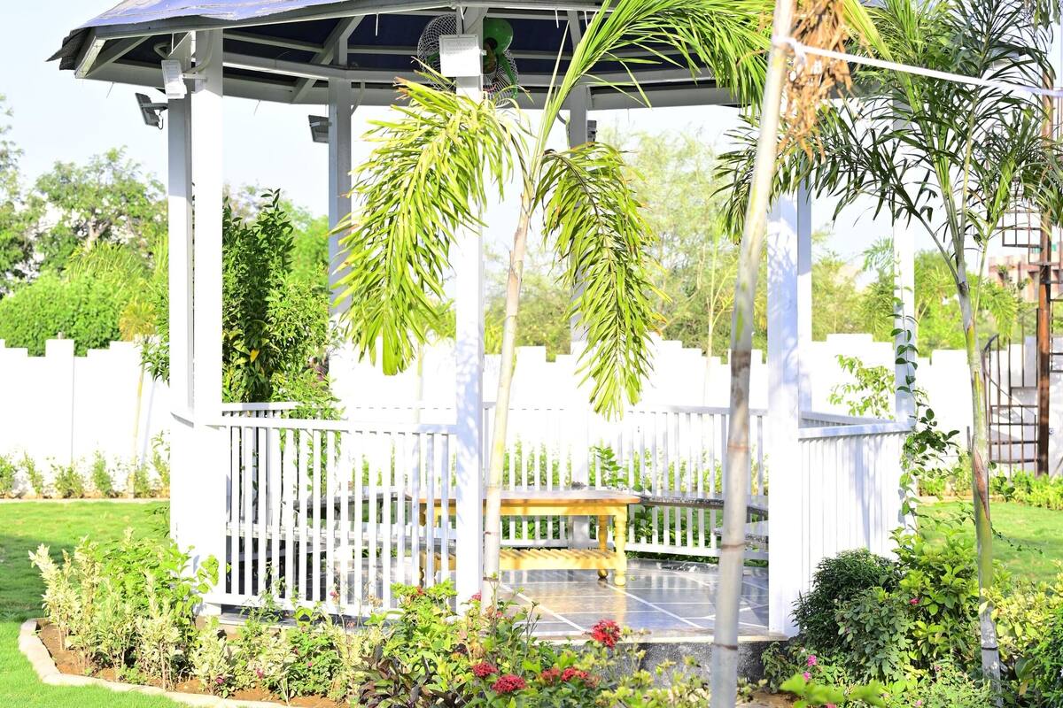 A gazebo with a blue roof, surrounded by a white fence and lush greenery, including palm trees and flowering plants.