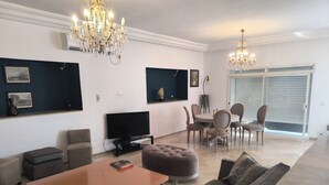 Een moderne woonkamer met een flatscreen televisie, een houten salontafel, een bank en een eethoek met stoelen.