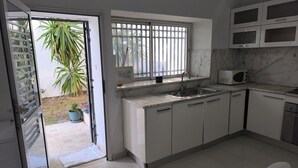Een moderne keuken met witte kastjes, een spoelbak en een magnetron. Er is een raam met tralies, en buiten staat een potplant zichtbaar.