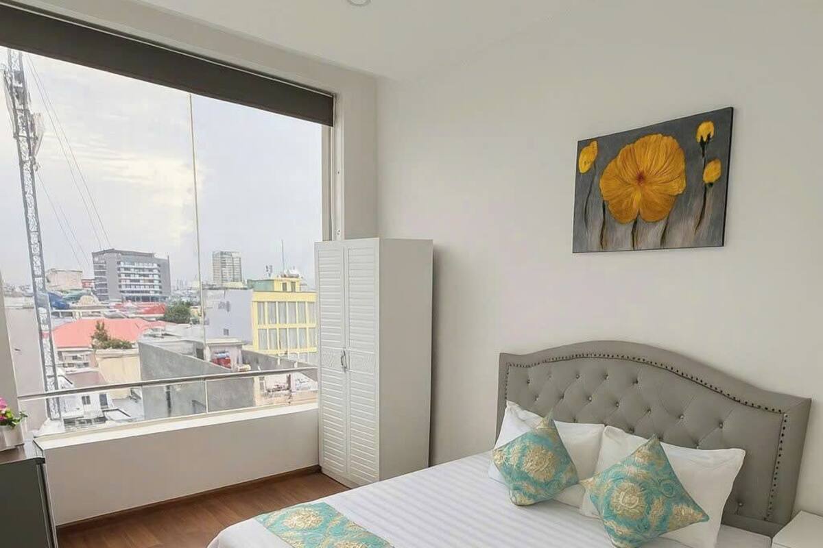 Une chambre avec une grande fenêtre, un lit doté d’une tête de lit capitonnée et un tableau représentant des fleurs accroché au mur.
