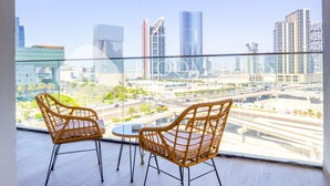 Un balcón con dos sillas de mimbre y una mesita, con vistas a un paisaje urbano de edificios modernos y una calle concurrida.