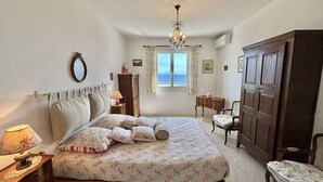 Une chambre à coucher comprenant un lit, une table de chevet, une commode, une chaise, un bureau, un lustre et une fenêtre donnant sur un beau paysage.