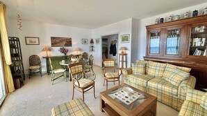 Un salon avec un canapé à carreaux, un pouf assorti et une table basse en verre. On y trouve également un coin repas avec une table et des chaises, ainsi qu’une bibliothèque remplie de livres.