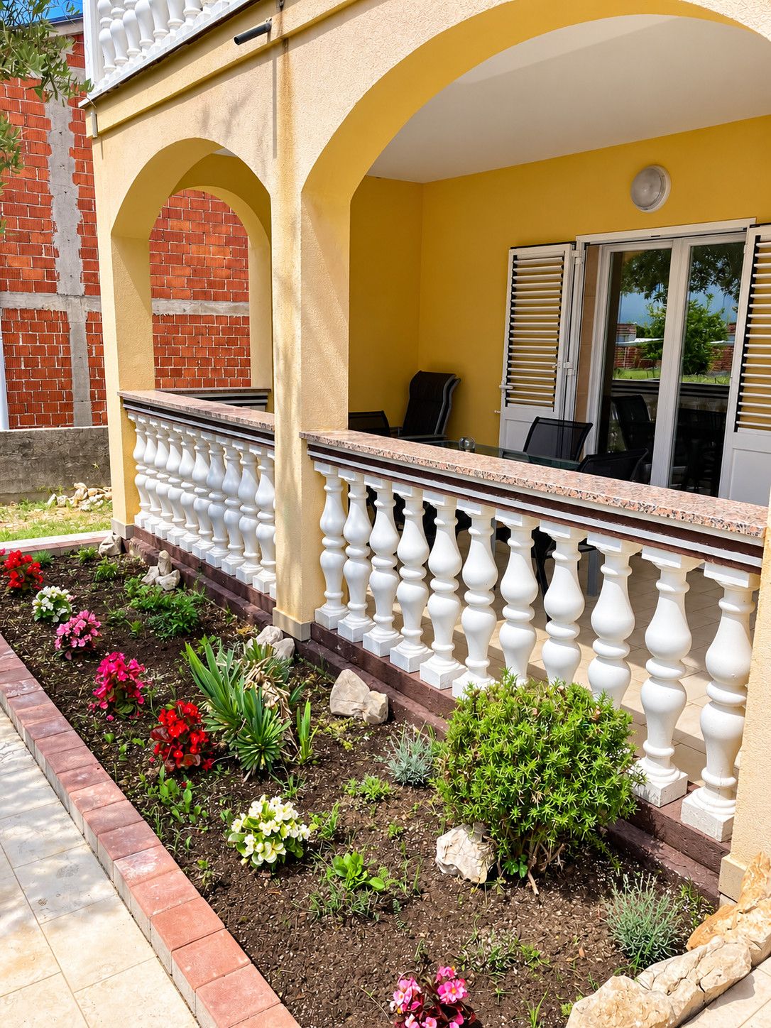 Een geel gebouw met een balkon, een bakstenen muur en een tuin met verschillende planten.