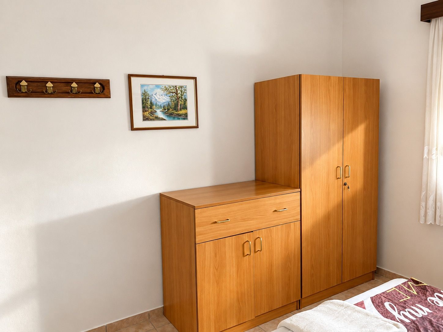 Une chambre à coucher avec une armoire en bois, une table de chevet, un tableau encadré au mur et une fenêtre avec des rideaux.