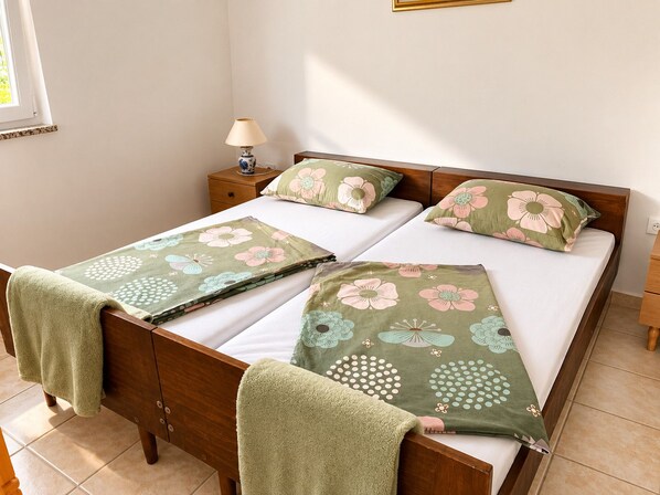 Quarto com cama de madeira, duas almofadas estampadas com flores, mesa de cabeceira com abajur e janela com persianas.