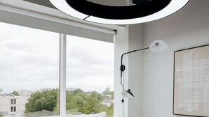 A modern hotel room with a large bed, a circular ceiling light, a window with a view of buildings and trees, and a wall-mounted lamp. - Chic 2BR/2BA Stay | Downtown & Capitol Views (Montgomery)