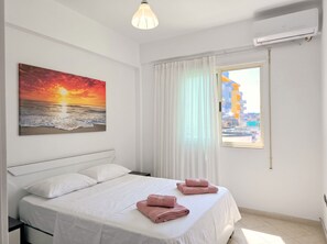 A hotel room with a bed, a painting of a sunset, a window with curtains, and an air conditioning unit. - Mandalay Suites by the Beach 079 (Durres)