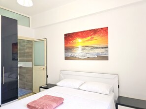 A hotel room with a bed, a nightstand, a painting of a sunset over the ocean, and a bathroom with a glass door. - Mandalay Suites by the Beach 079 (Durres)