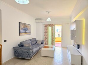 A modern living room with a grey sofa, a white coffee table, a painting, and a TV on the wall. - Mandalay Suites by the Beach 079 (Durres)