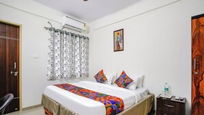 Una camera d'albergo con un letto, un comodino, una porta in legno, una finestra con tende a motivi e un condizionatore d'aria.