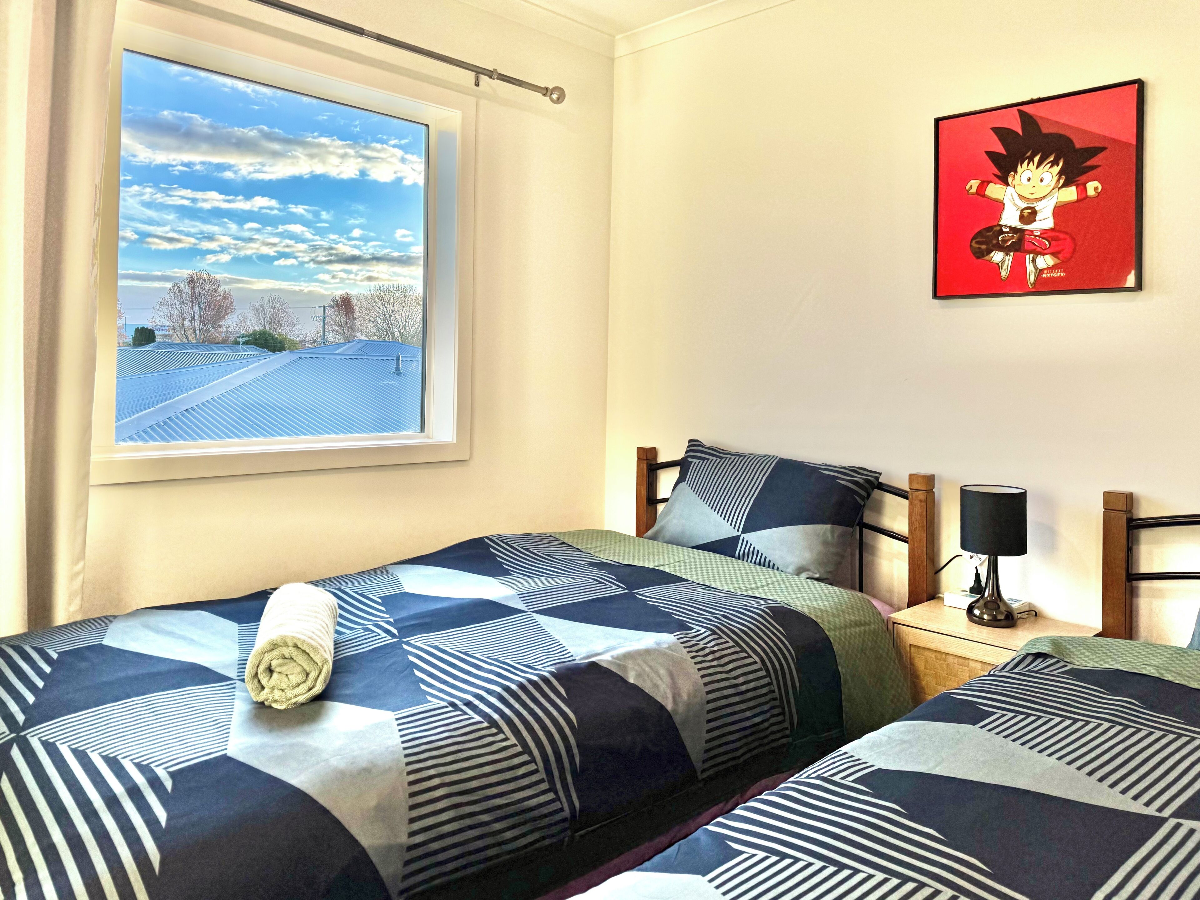 Een slaapkamer met twee bedden, een raam met uitzicht op een blauwe lucht en bomen, een wandlamp en een ingelijst schilderij.