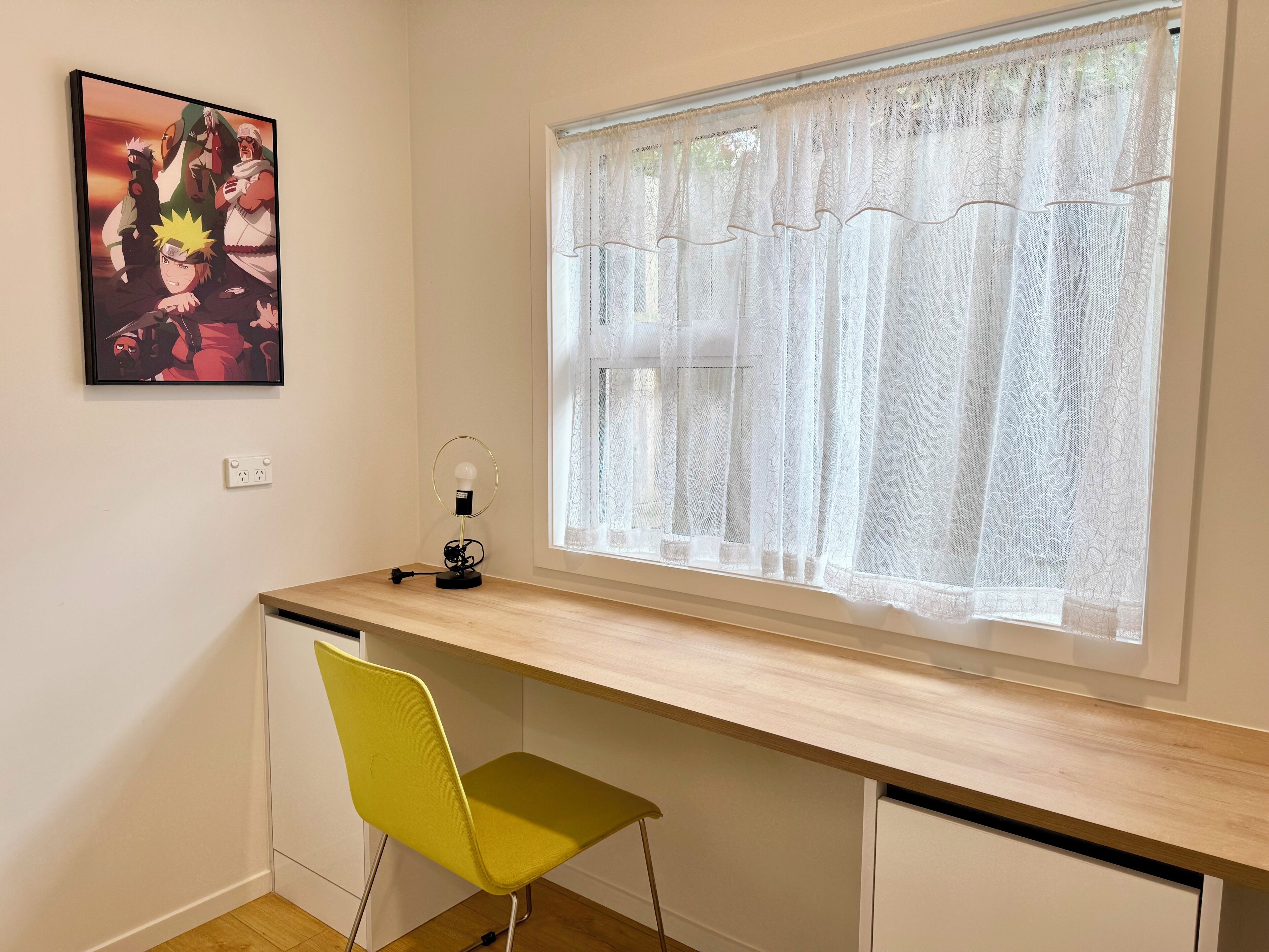 A room with a desk, a chair, a window with lace curtains, and a framed picture on the wall.