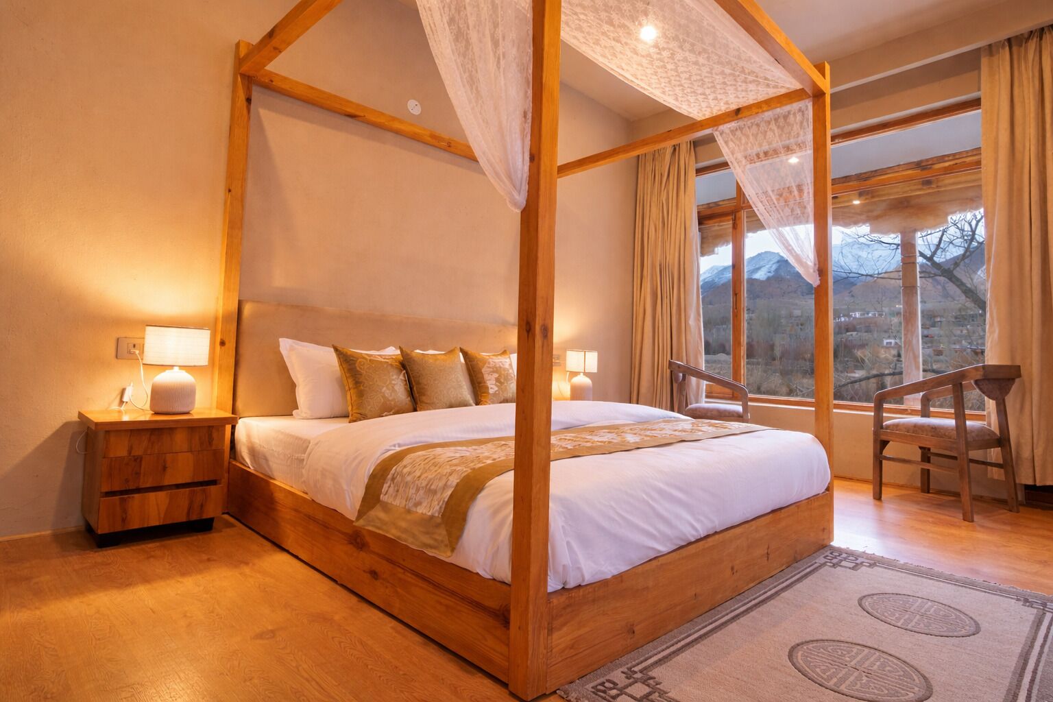 Una cama con dosel, mesitas de noche de madera, una silla y vistas a montañas a través de ventanas amplias.