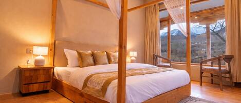 Una cama con dosel, mesitas de noche de madera, una silla y vistas a montañas a través de ventanas amplias.