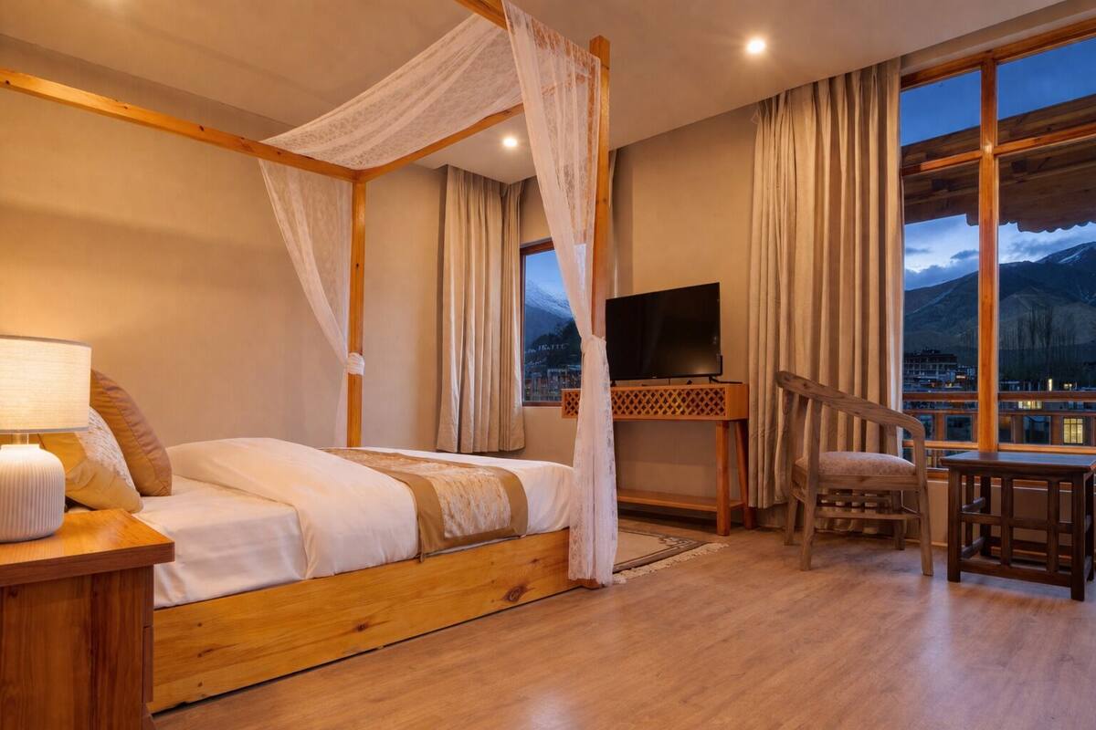 A bedroom with a four-poster bed, a TV on a wooden stand, a chair, and a view of mountains through large windows.