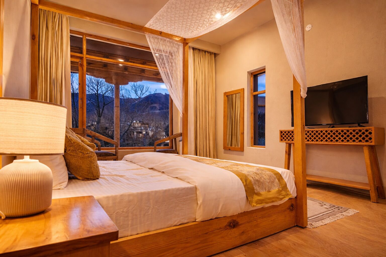 A bedroom with a bed, a bedside table, a lamp, a TV, and a view of the outside.