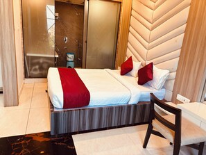 Una habitación de hotel con cama, una silla de madera, una mesita pequeña y zona de ducha.