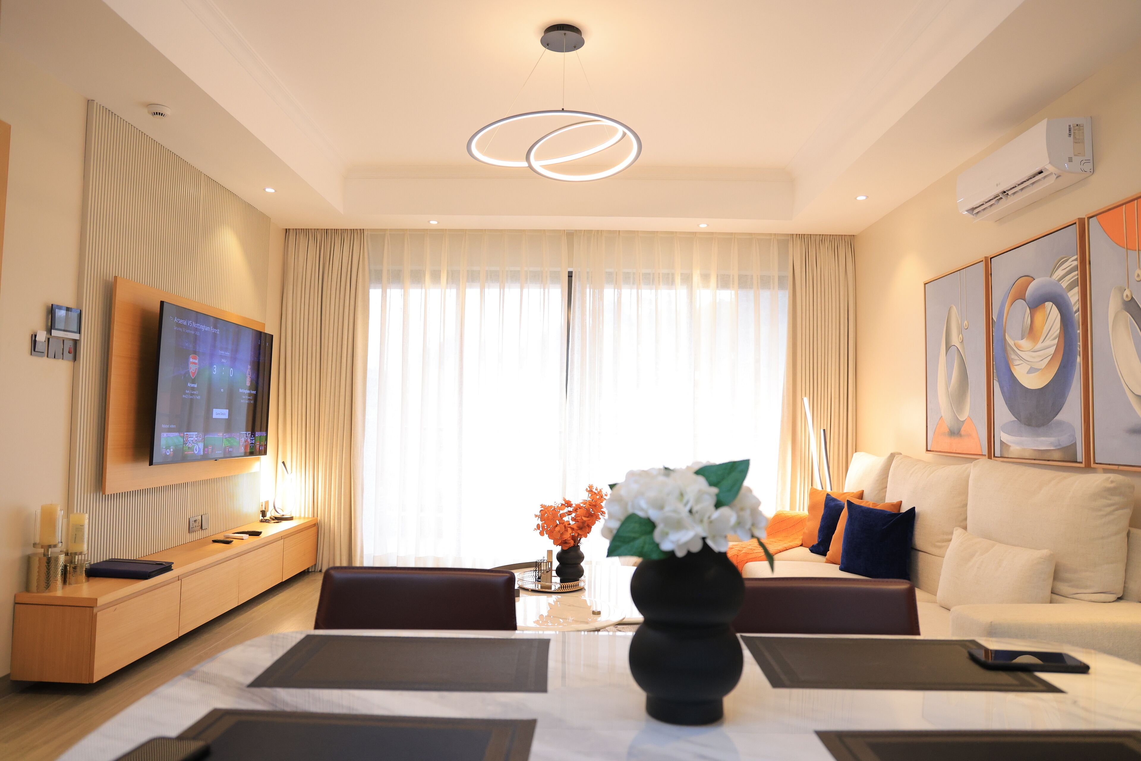 A modern hotel room with a flat-screen TV, a dining table with a vase of flowers, a sofa, and artwork on the wall.