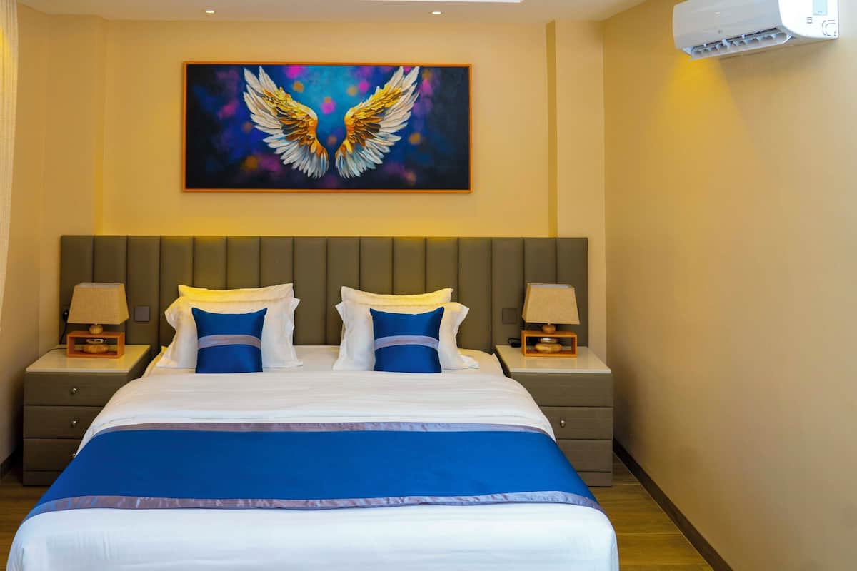 A hotel room with a large bed, two bedside tables with lamps, a painting of wings on the wall, and an air conditioning unit.