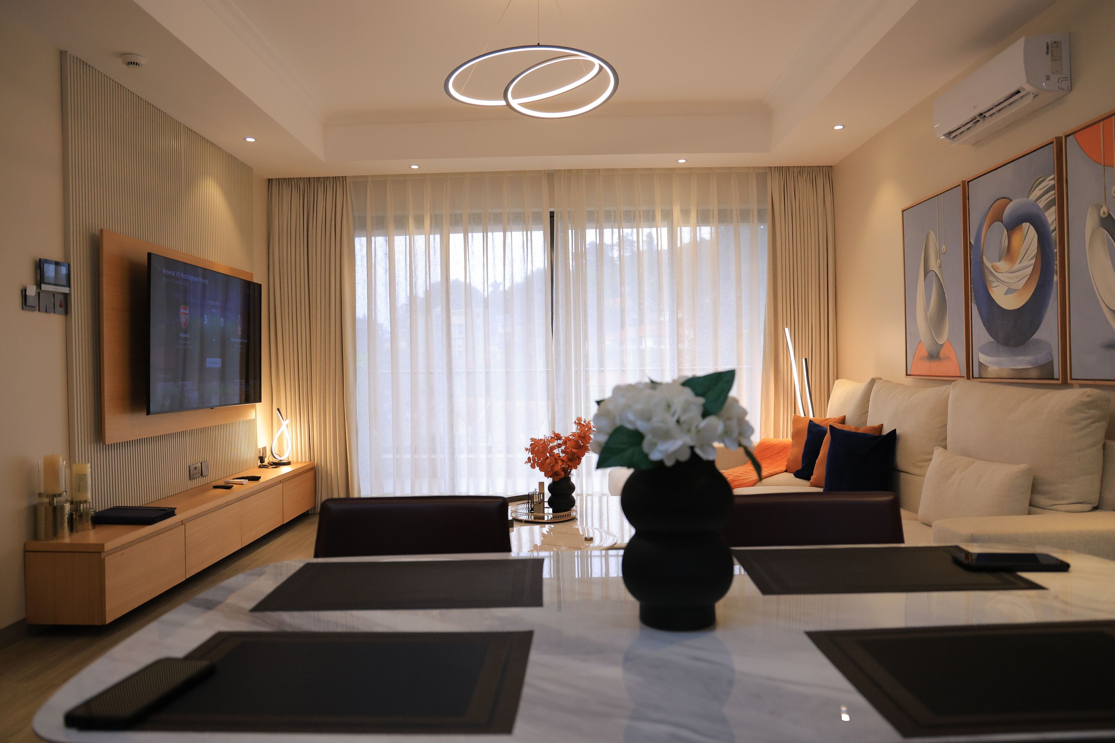 A modern hotel room with a flat-screen TV mounted on the wall, a sofa, a dining table with placemats, and a vase with flowers.