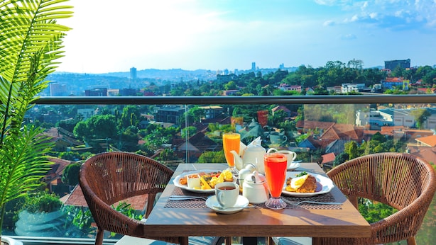 A balcony with wicker chairs, a table with breakfast, and a city view. - Abrish Hotel and Apartment (Kampala)