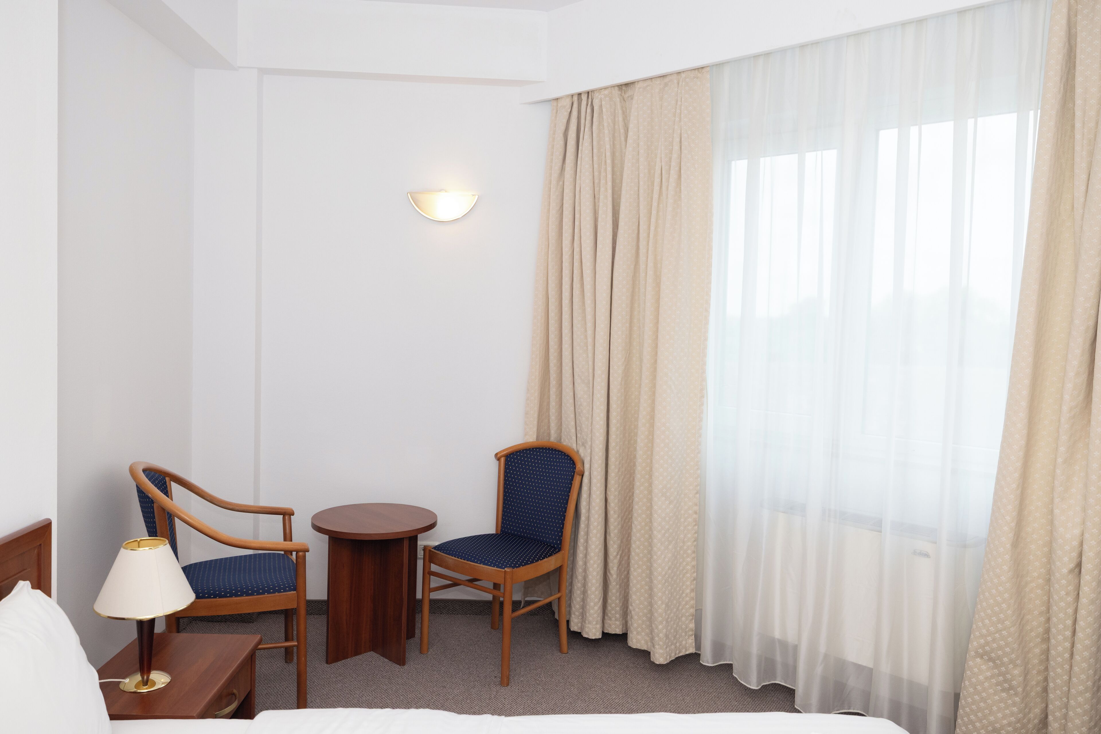 Une chambre d’hôtel avec un lit, deux chaises, une petite table, une lampe et une fenêtre avec des rideaux légers.