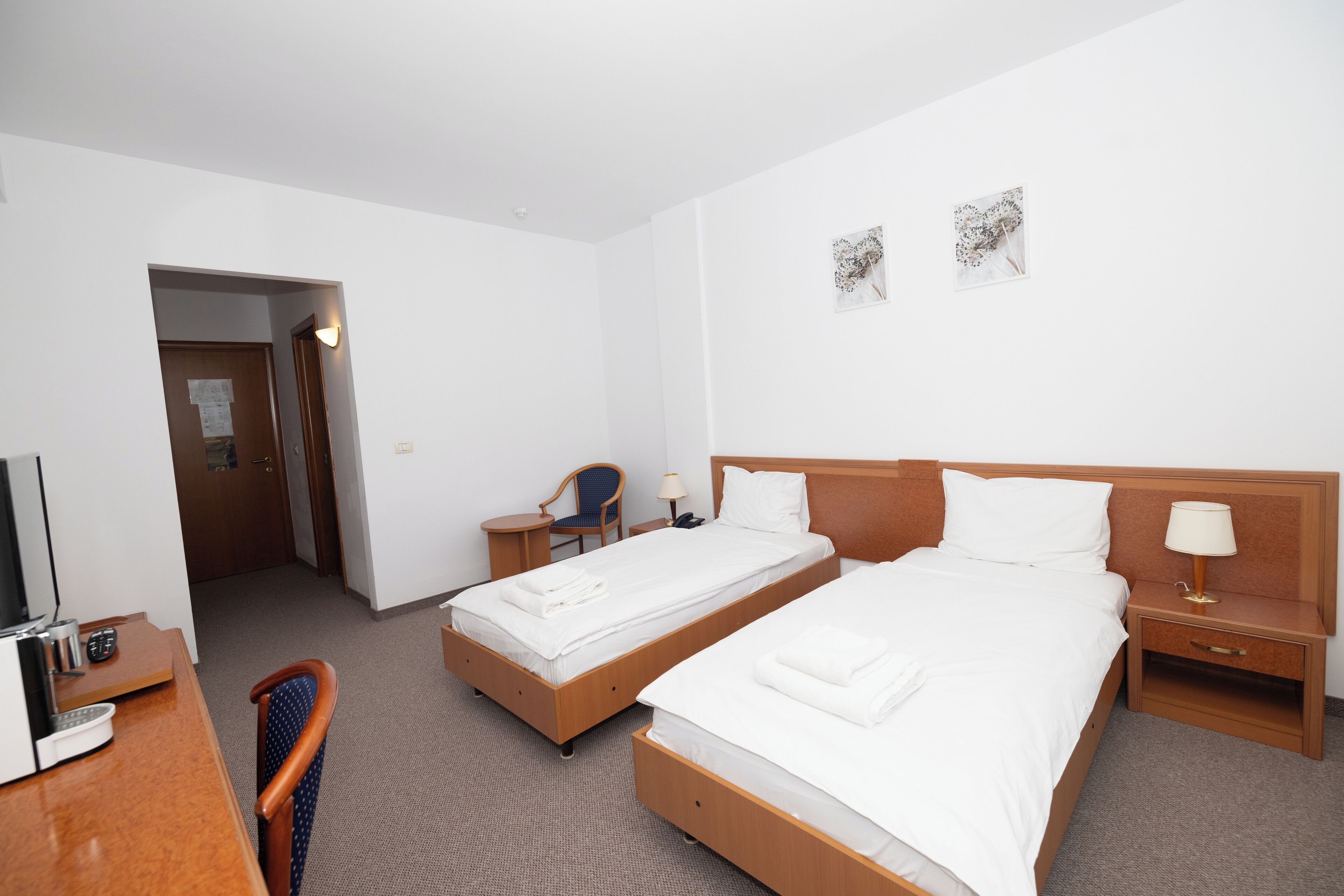 A hotel room with two single beds, a desk with a chair, a small table, and a nightstand with a lamp.