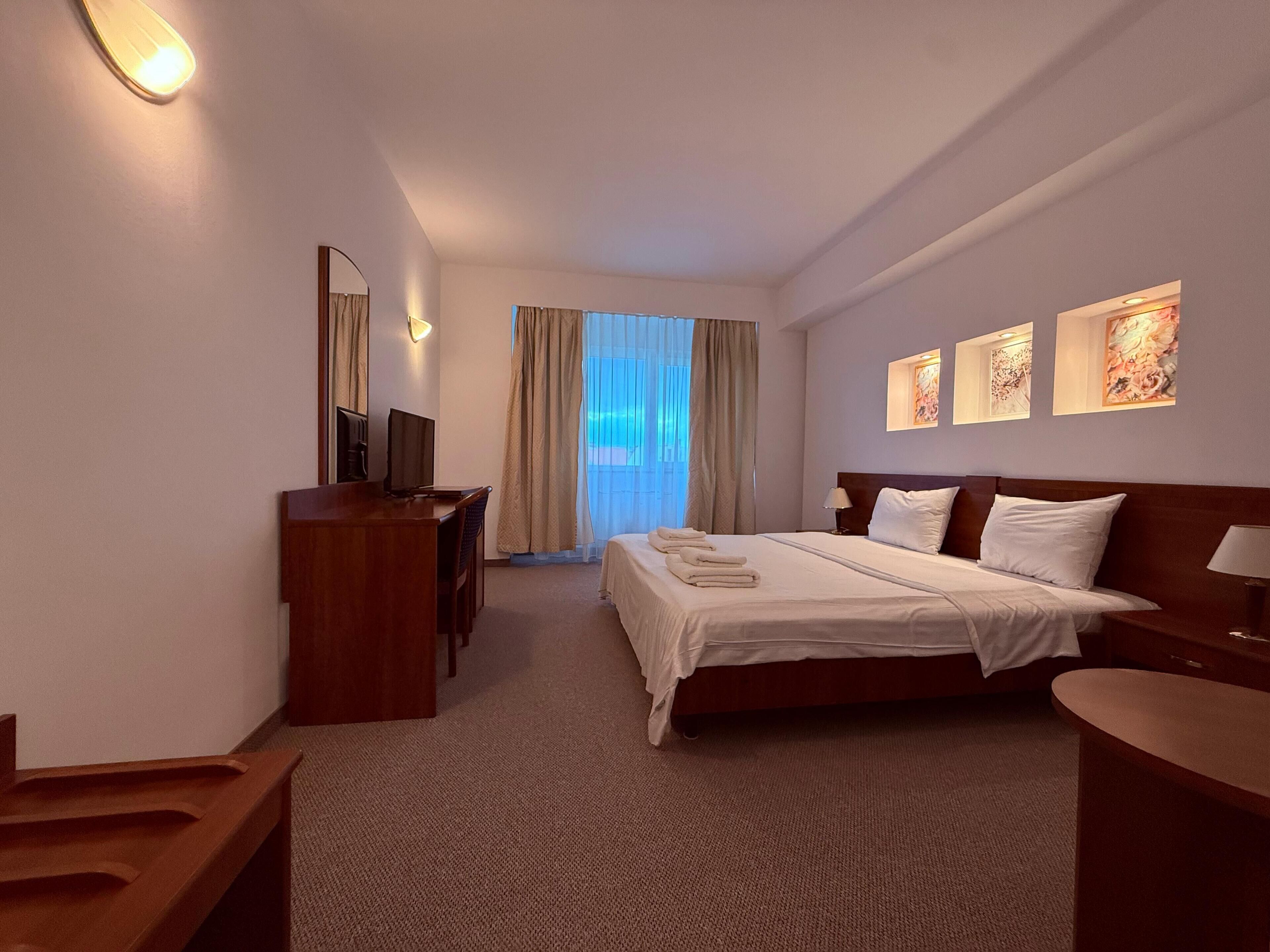 Une chambre d’hôtel avec un lit, un bureau, une chaise, une télévision et une fenêtre avec des rideaux.