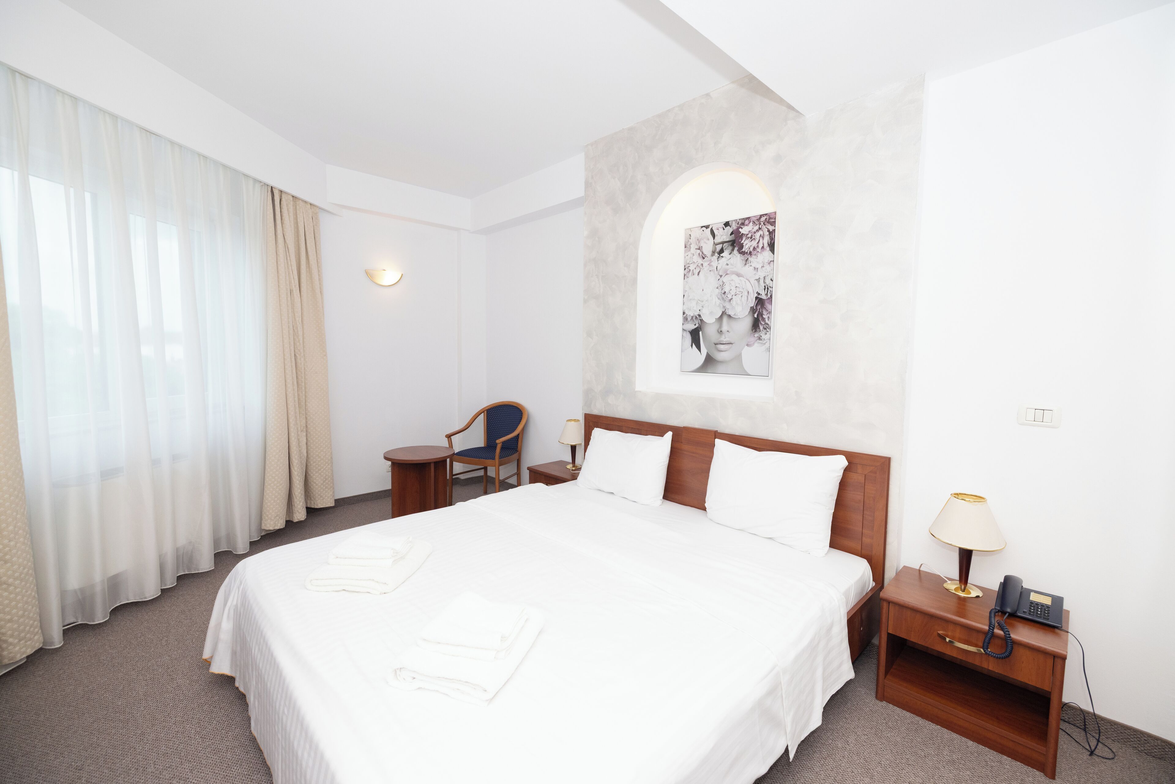 Habitación de hotel con una cama grande, una silla, una mesita de noche con lámpara y teléfono, y un cuadro colgado en la pared.