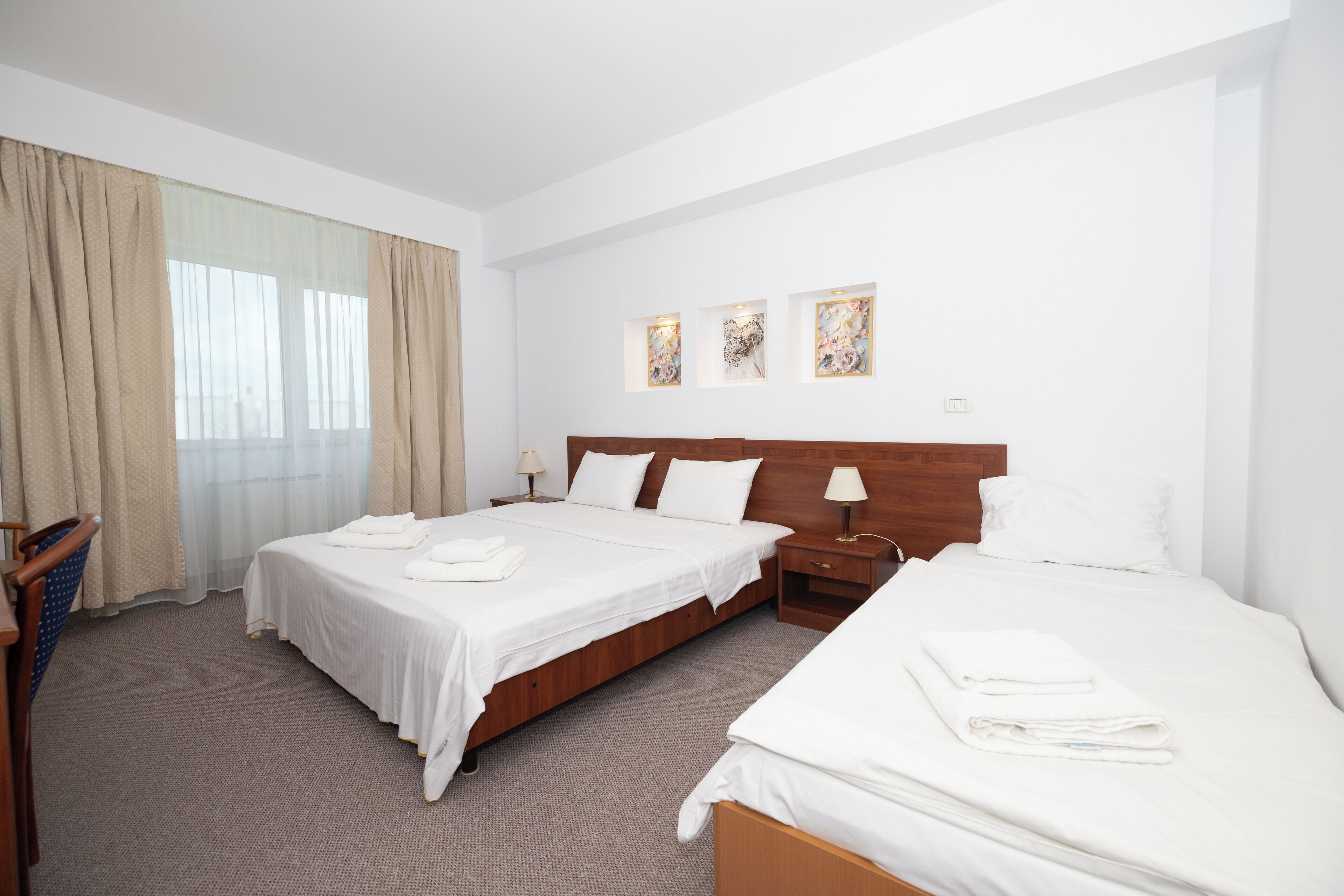 A hotel room with two beds, a wooden headboard, a large window with curtains, and three framed artworks on the wall.