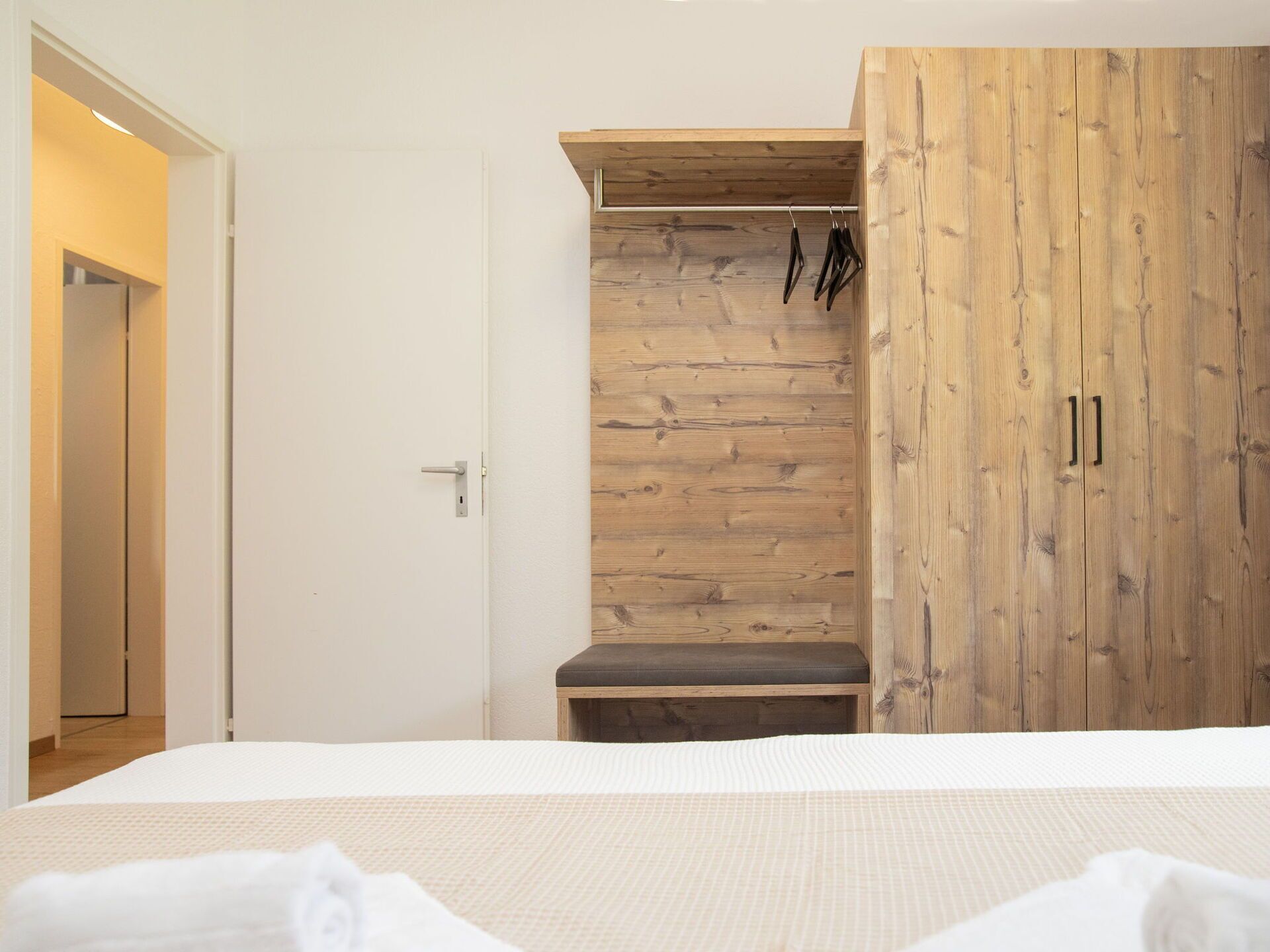 A bedroom with a wooden wardrobe, a bench, and a bed with a white duvet.